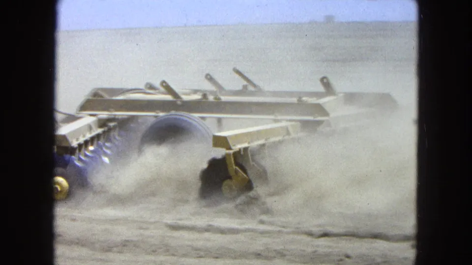 Close 8mm view of a tractor-drawn plow or harrow cutting through dry soil with dust rising around the metal frame.