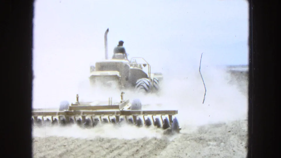 Rear view of a tractor operator tilling a dry Pueblo, Mexico field, with a wide implement and dust filling the 8mm frame.