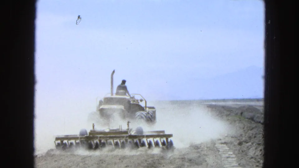 A farmer on a tractor pulls a wide disc harrow through a dry field, with haze and distant hills visible in the 1965 Mexico footage.