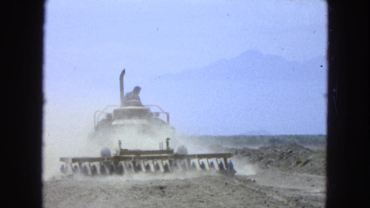 Tractor with front blades moves across dusty dry ground with a seated operator.