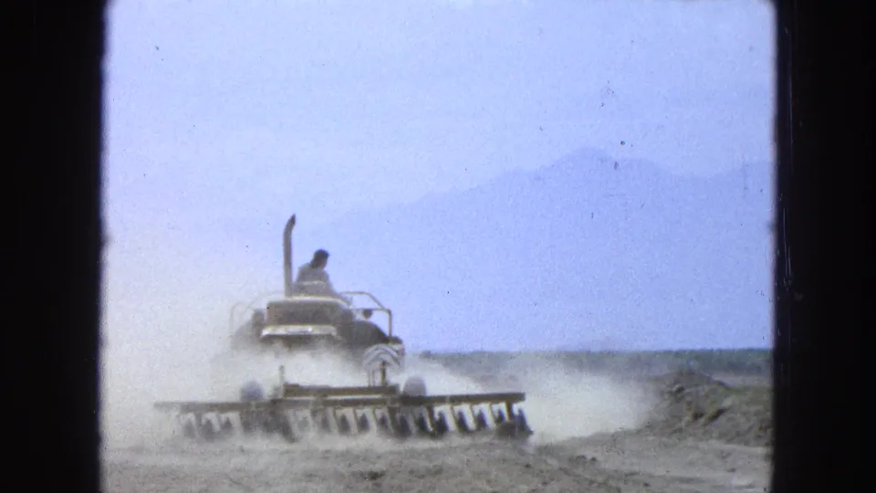 A wide 8mm frame shows the tractor working dry soil, with the operator visible and mountains softened by haze.