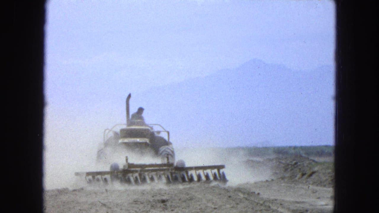 Tractor operator drives through a dusty field with a plow attachment and distant mountains behind.