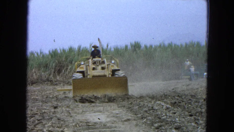 1965 Pueblo, Mexico 8mm frame showing a bulldozer pushing loose soil in a dusty field, with cane behind and a distant figure near the right edge.
