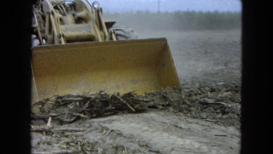 1965 8mm frame from Pueblo, Mexico showing a yellow tractor blade moving soil and plant debris in an open field.