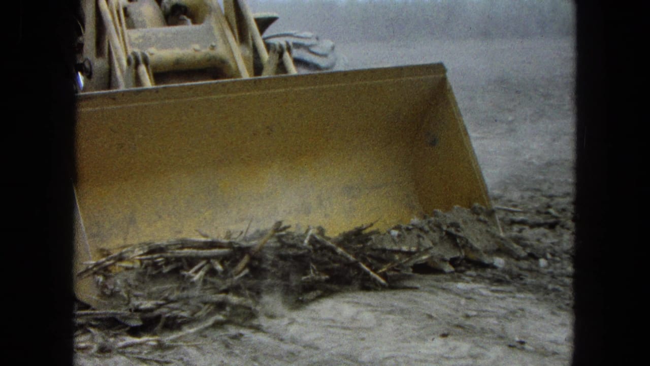Close 8mm view of a yellow tractor blade leveling soil and crop residue in Pueblo, Mexico, in 1965.
