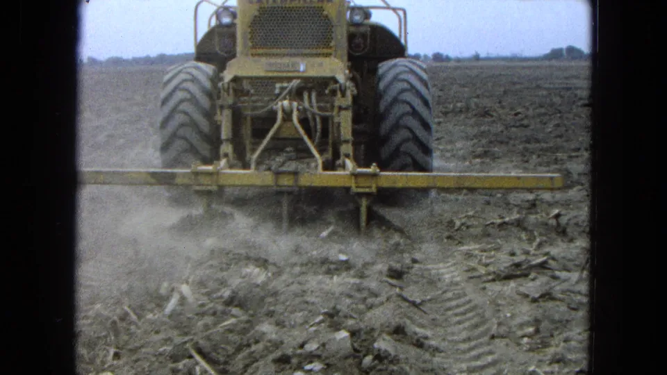 Rear view of a yellow tractor with large tires and leveling equipment moving through a dusty farm field in 1965 Pueblo, Mexico.