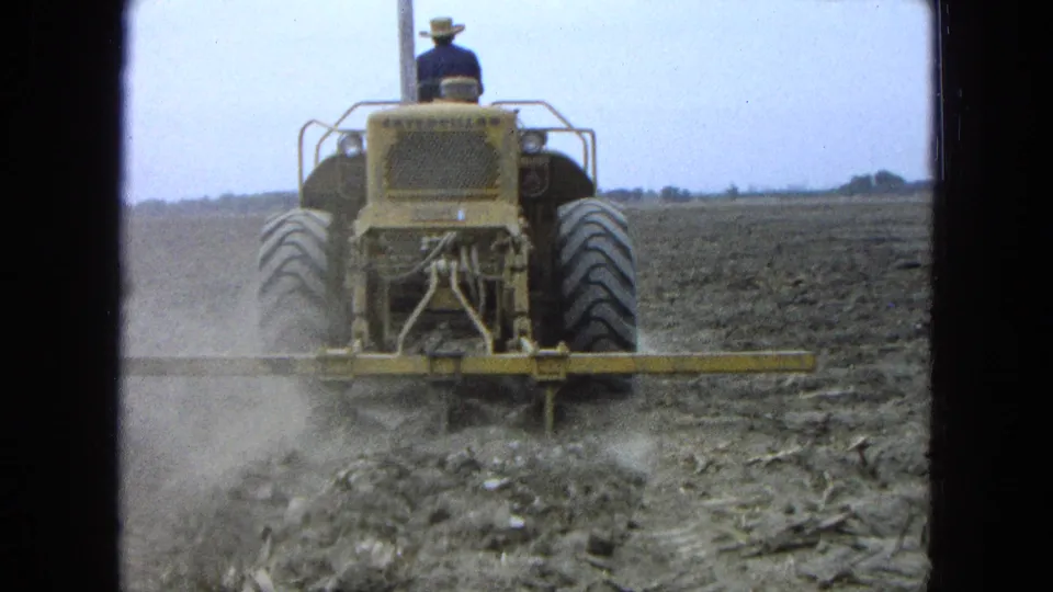 A driver in a wide-brimmed hat operates a yellow tractor through a dusty field in 1965 Pueblo, Mexico.