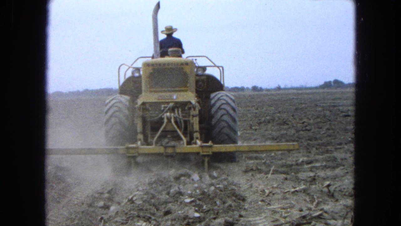 Farmer drives a yellow tractor away from the camera across a dusty plowed field.