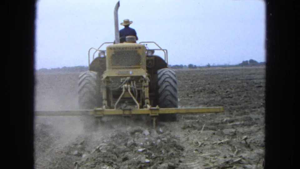 1965 Pueblo, Mexico footage shows a farmer in a straw hat driving a yellow tractor over rough tilled soil.