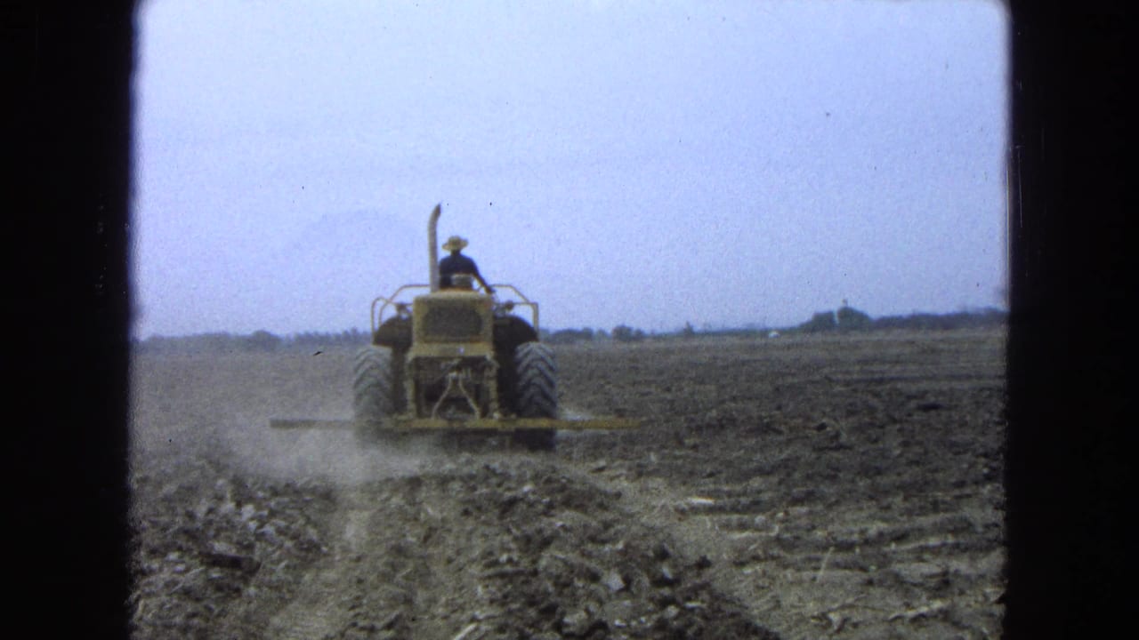 Yellow tractor moves farther across the field, leaving dust over the tilled rows.