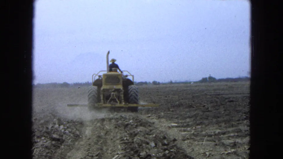 A farmer drives a yellow tractor away across a broad Pueblo, Mexico field, with dust trailing behind the plow.