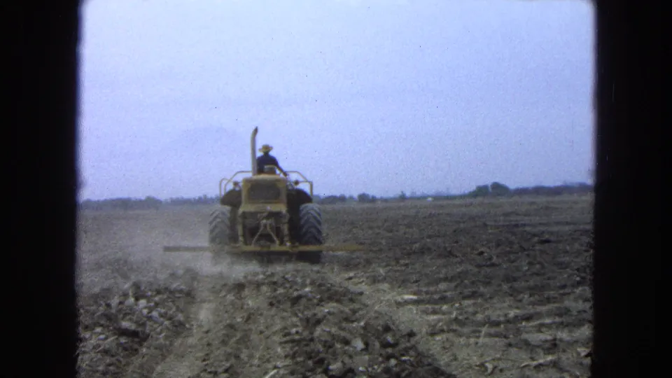The 8mm frame shows the tractor receding across the dark tilled field under a pale overcast sky.