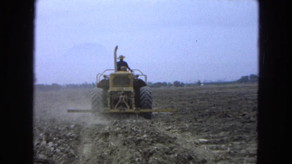 A rear view of the yellow tractor shows continued plowing in a broad rural field in the 1965 Mexico clip.