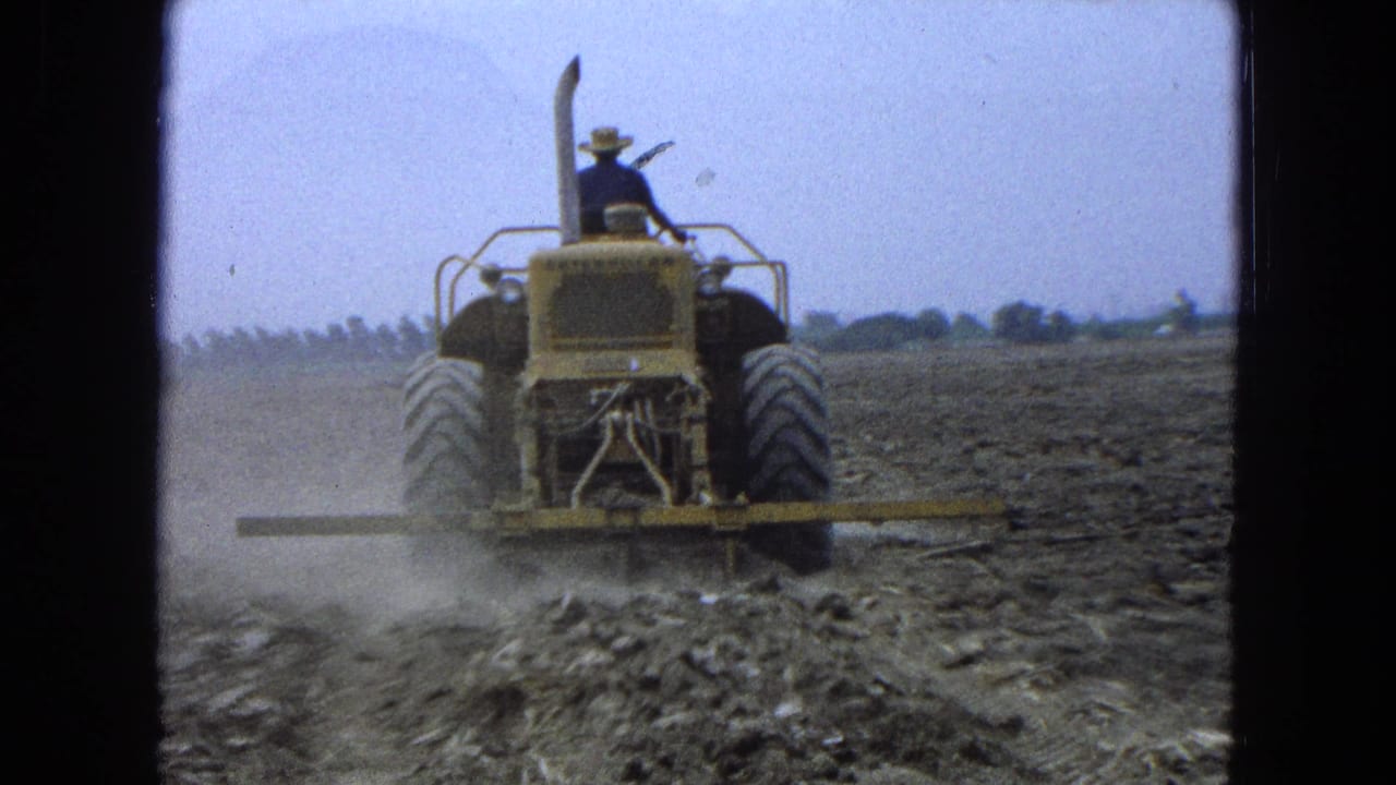 Farmer in a hat drives a yellow tractor across a dusty field.