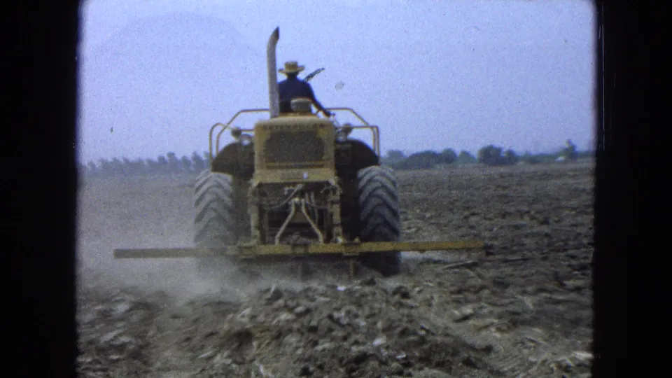 In Pueblo Mexico, 1965, a farmer in a hat drives a yellow tractor through dry farmland as dust rises behind it.