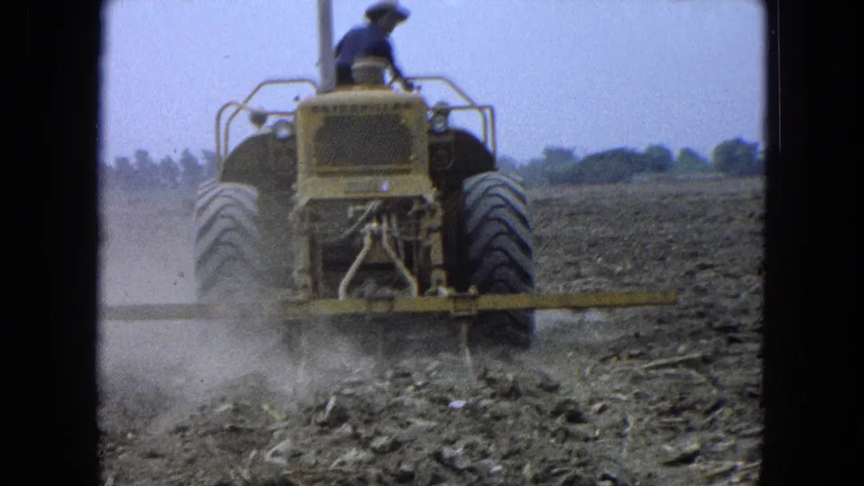 The 1965 8mm frame shows a farmer on a yellow tractor crossing dry farmland in Pueblo Mexico, with dust near the wheels.