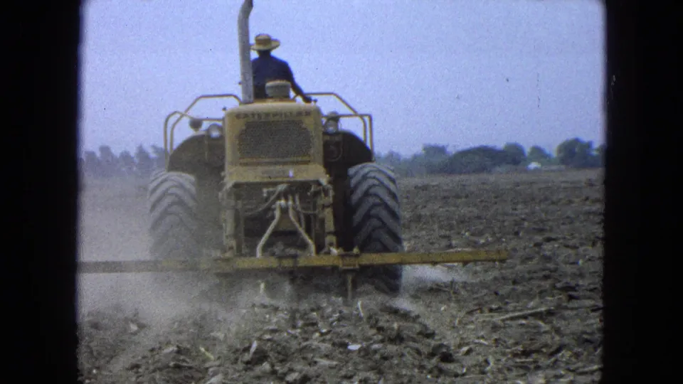 A farmer in a hat drives a yellow tractor away through dry Pueblo Mexico farmland, filmed on 8mm in 1965.