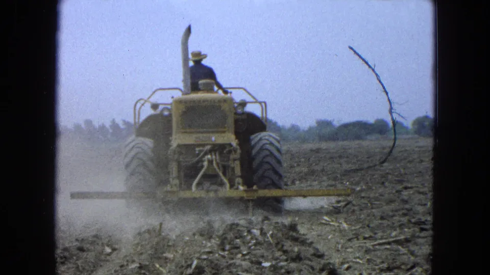 A straw-hatted farmer drives the tractor away from the camera while dust hangs over the worked soil and a film scratch marks the 8mm frame.