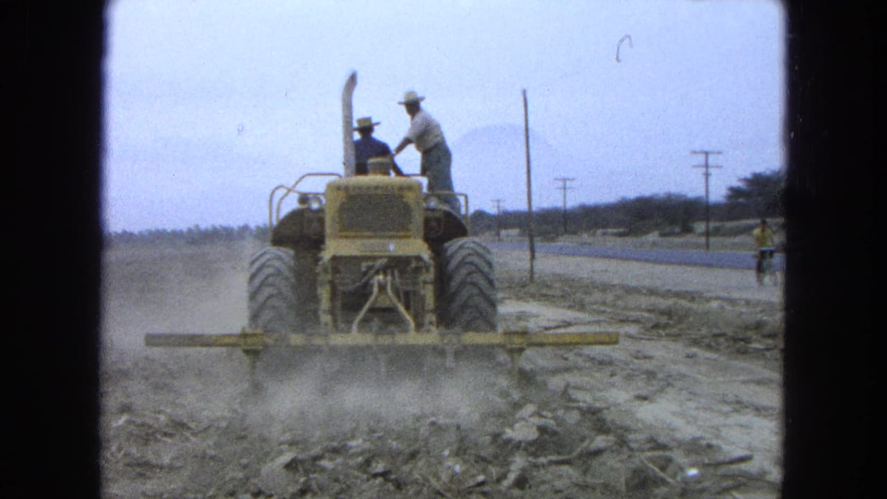Two men ride a yellow tractor across a dry field beside a road.