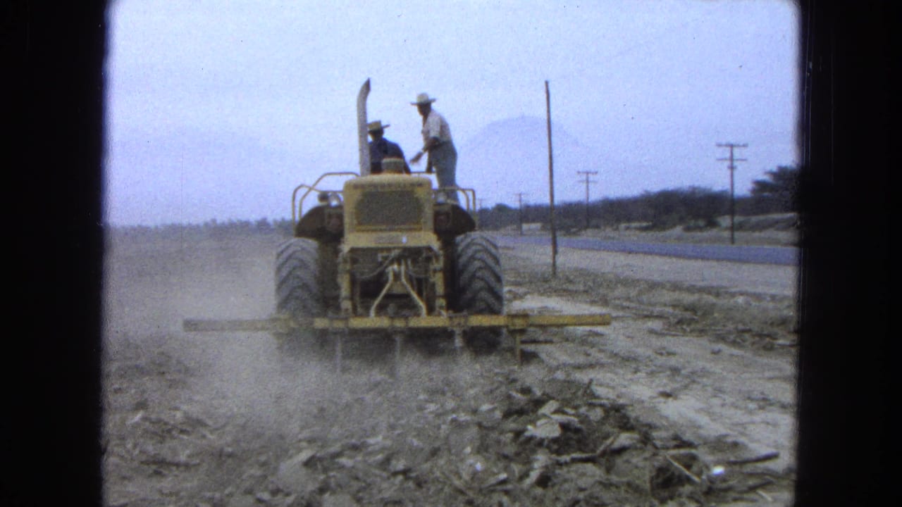 Yellow tractor travels away through dust with two men on board.