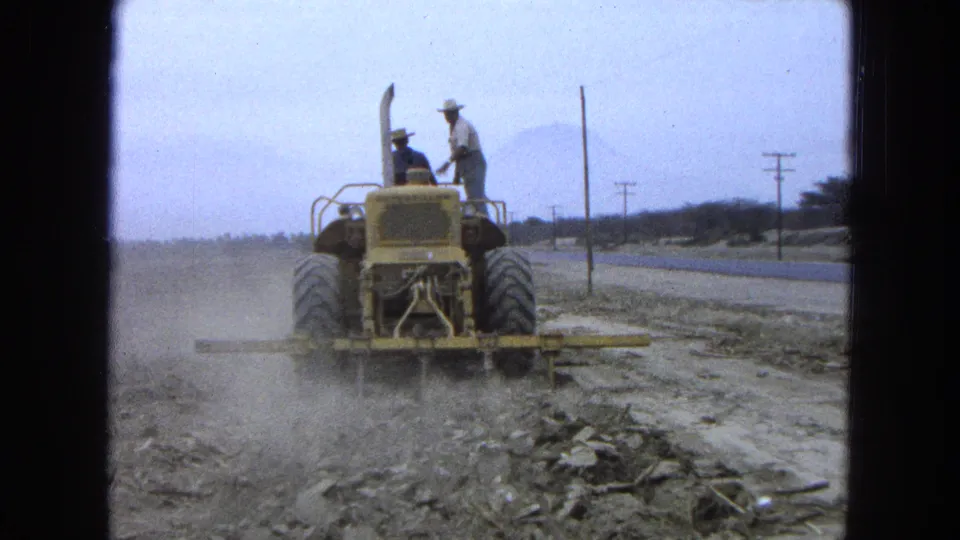 The tractor continues across the field in Pueblo, Mexico, kicking up dust as two men ride on its rear platform.