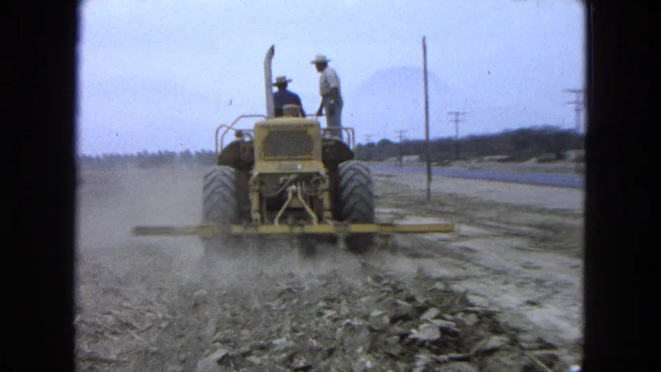A rear view shows the yellow tractor working through the dry field, with two men visible against the pale sky.