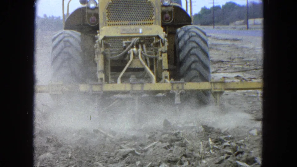Close rear framing shows the tractor's large tires, rear grille, and field equipment stirring dust in the 1965 rural Mexico scene.