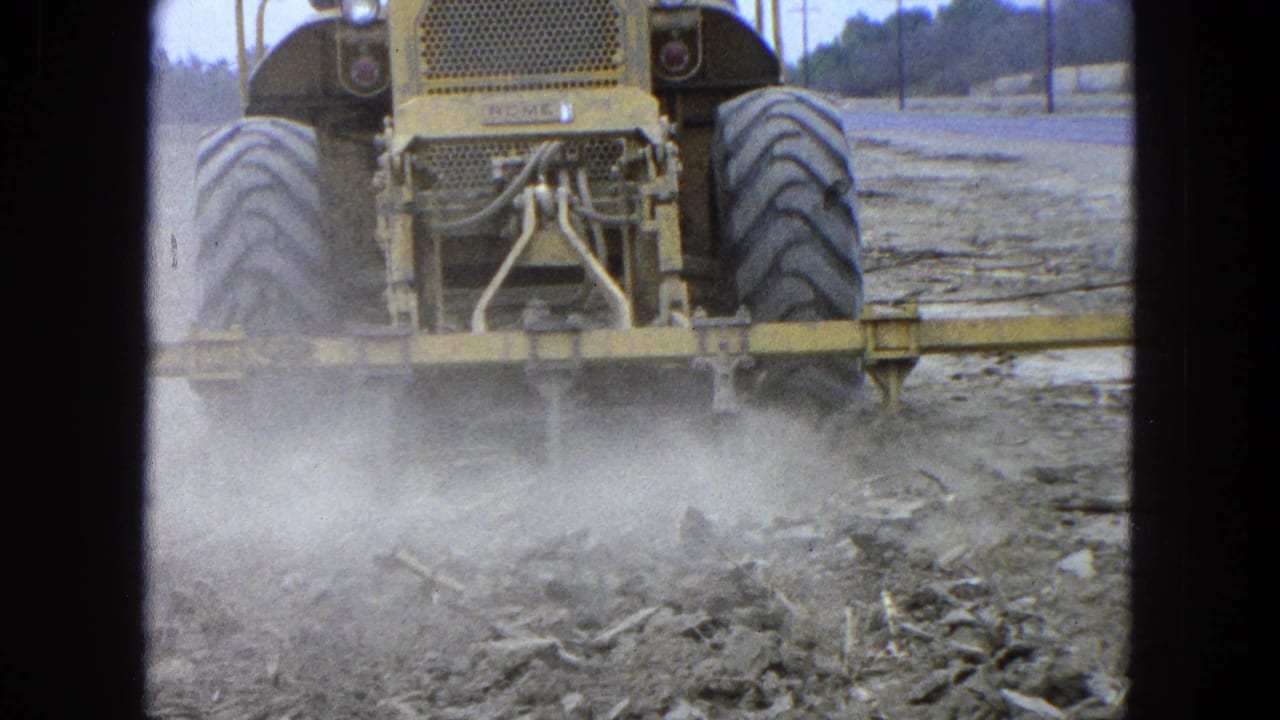 Rear view of a yellow tractor kicking dust while tilling a dry field.