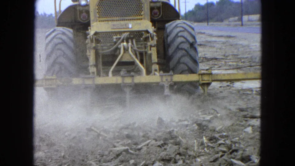 1965 Pueblo Mexico 8mm frame showing a yellow tractor from behind as its tillage attachment churns dusty soil.