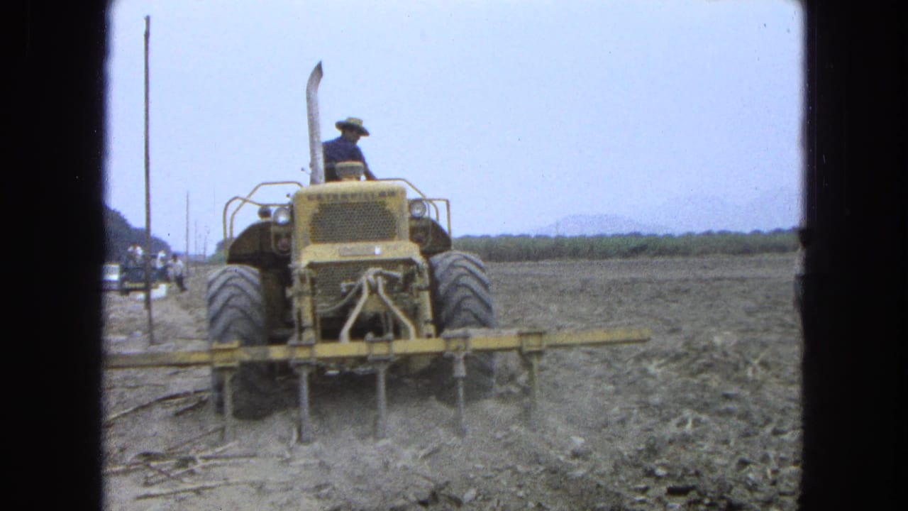 Tractor moves through a dry field near a road edge, with poles and distant hills behind it.