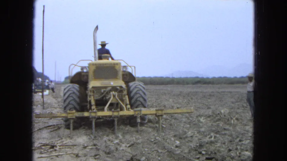 The 8mm frame shows a rear view of the yellow tractor and tilling bars working rough soil in a rural Pueblo, Mexico field.