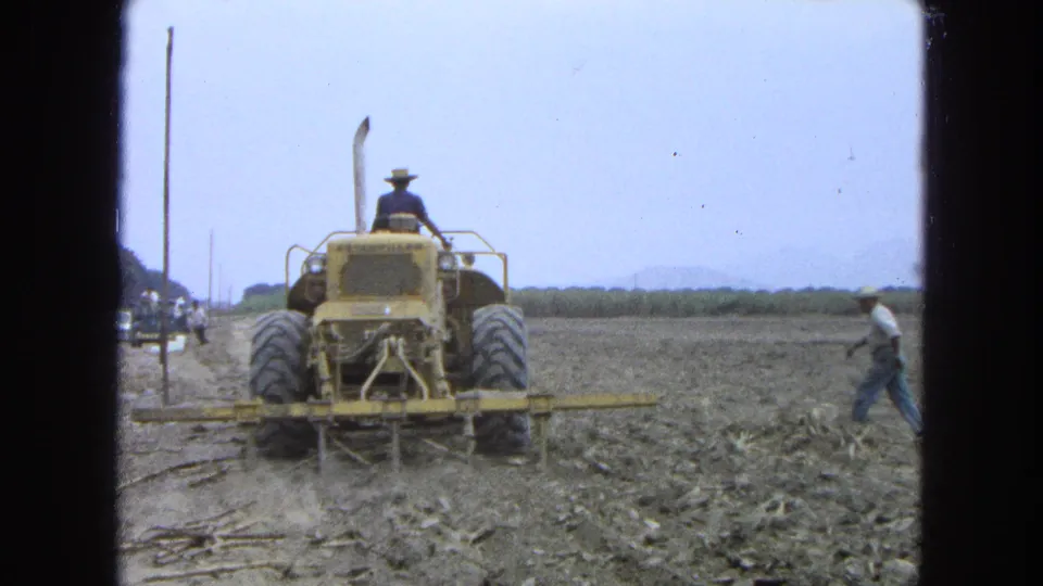 A yellow tractor continues field work in 1965 Pueblo, Mexico as another worker walks along the dry field edge at right.