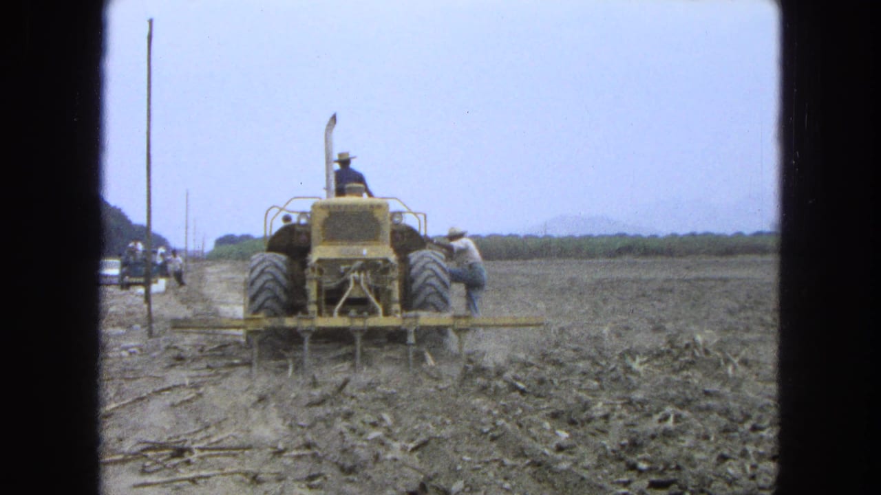 1965 Pueblo, Mexico frame with a yellow tractor plowing forward while a worker stands near the equipment in a dry field.