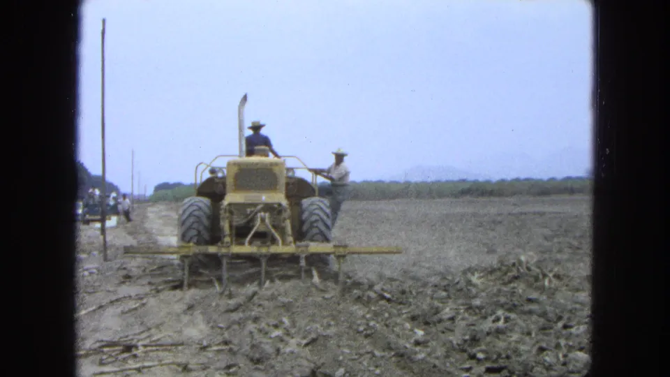 1965 Pueblo, Mexico 8mm frame showing the tractor and plow from behind, with a roadside vehicle and open field beyond.
