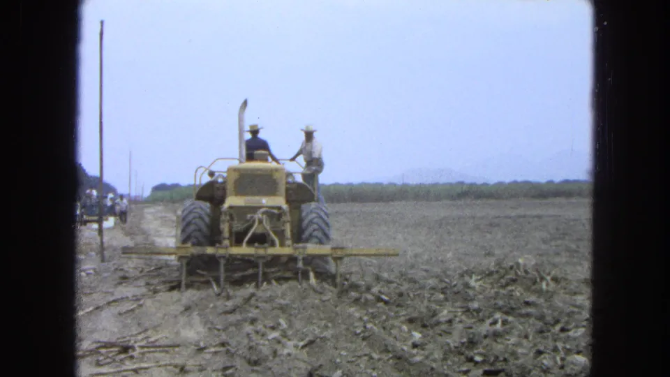 1965 Pueblo, Mexico frame showing two hat-wearing men around a yellow tractor as it works a dry rural field.