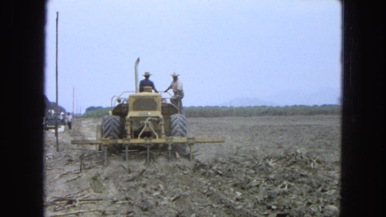 Yellow tractor drives through a dusty plowed field with two men riding and roadside figures in the distance.