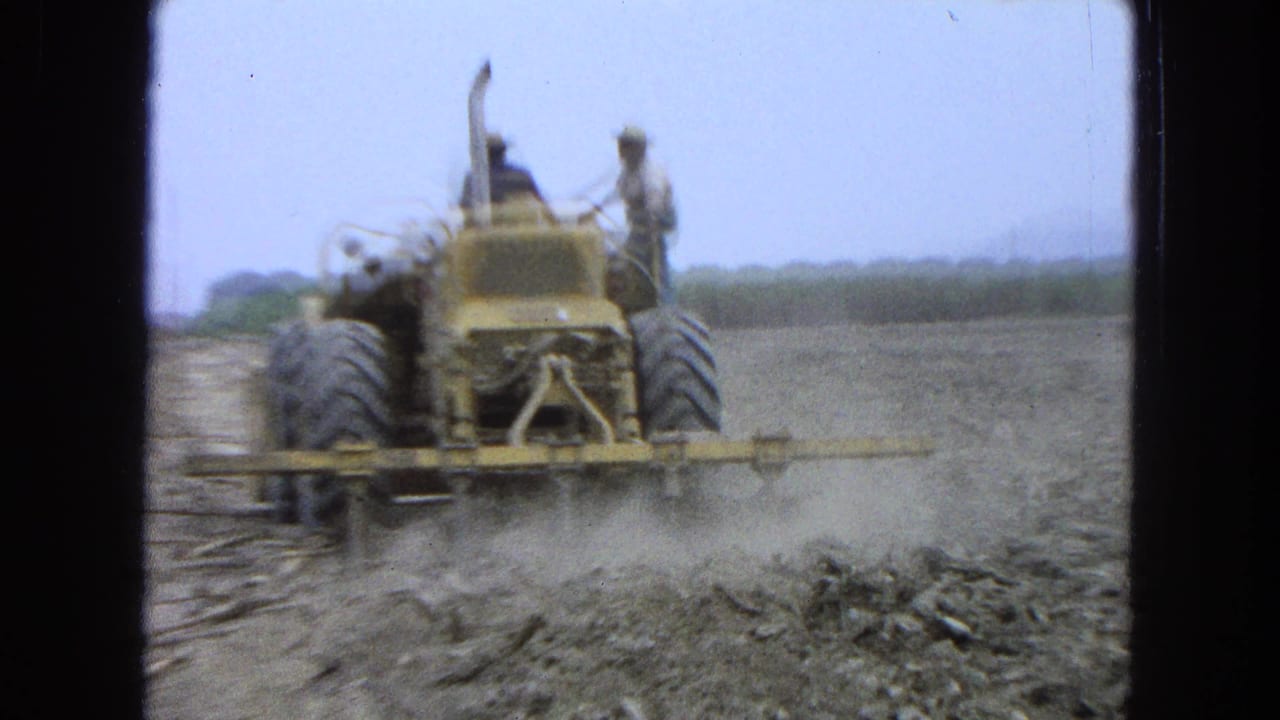 1965 Pueblo, Mexico 8mm frame with a rear view of a yellow tractor, two men aboard, and dust rising from field work in a dry rural landscape.