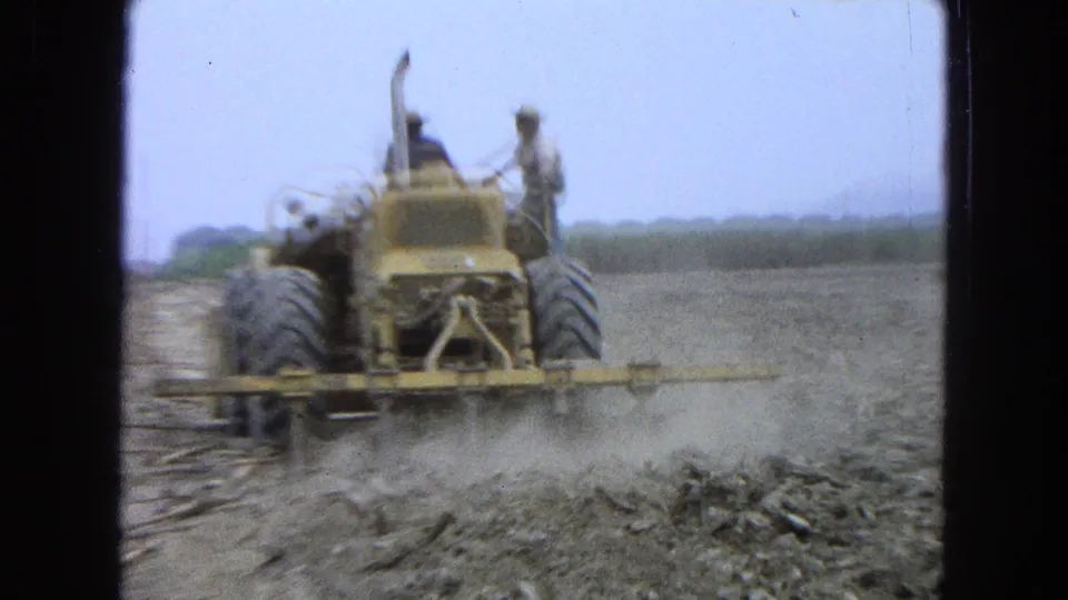 1965 Pueblo, Mexico 8mm frame with a rear view of a yellow tractor, two men aboard, and dust rising from field work in a dry rural landscape.