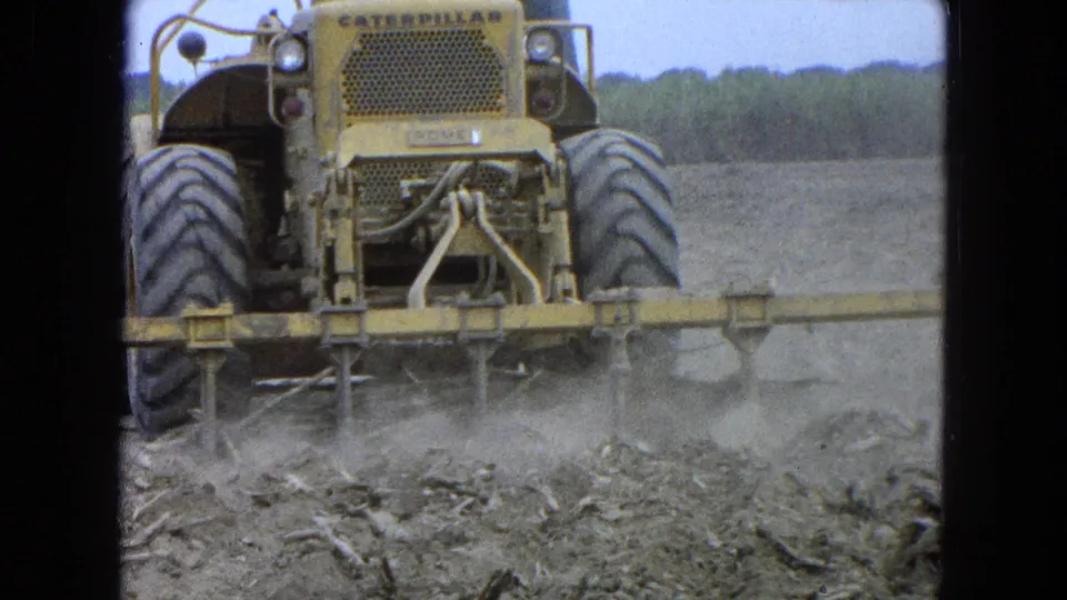 Close 1965 Pueblo, Mexico 8mm view of a yellow Caterpillar tractor, large rear wheels, and a low field attachment working dusty soil.