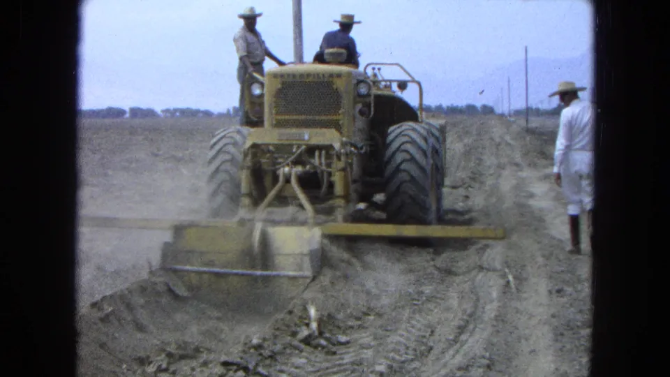 1965 Pueblo, Mexico 8mm frame showing a tractor working near a field road while two adult men ride and another man watches from the side.