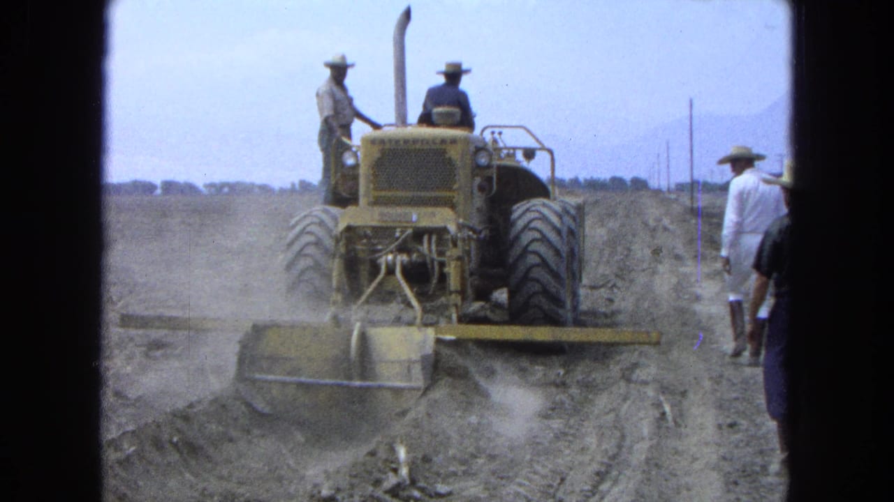 Yellow tractor leveling dusty dirt while men in hats walk beside it.