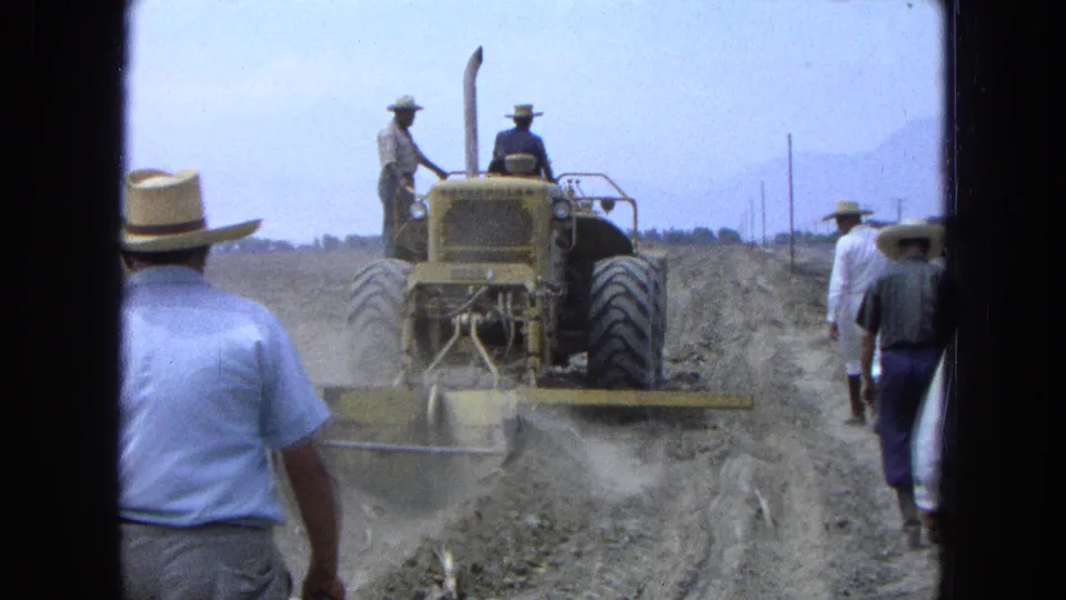 1965 Pueblo, Mexico view from behind a yellow tractor as workers in wide-brimmed hats walk along dusty graded ground.