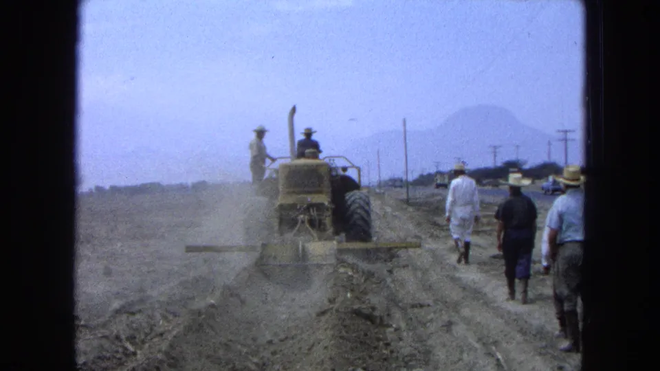 Wide 1965 Pueblo, Mexico frame of a yellow tractor moving away on a dusty path with workers walking behind and mountains faintly visible.