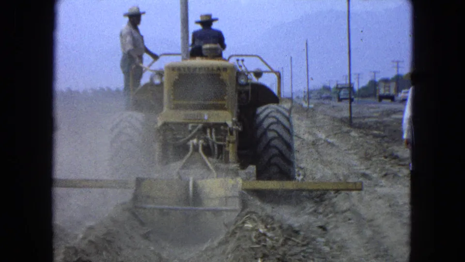 Close rear view from 1965 Pueblo, Mexico of a yellow tractor leveling rough dirt, with operators aboard and roadside poles beyond.