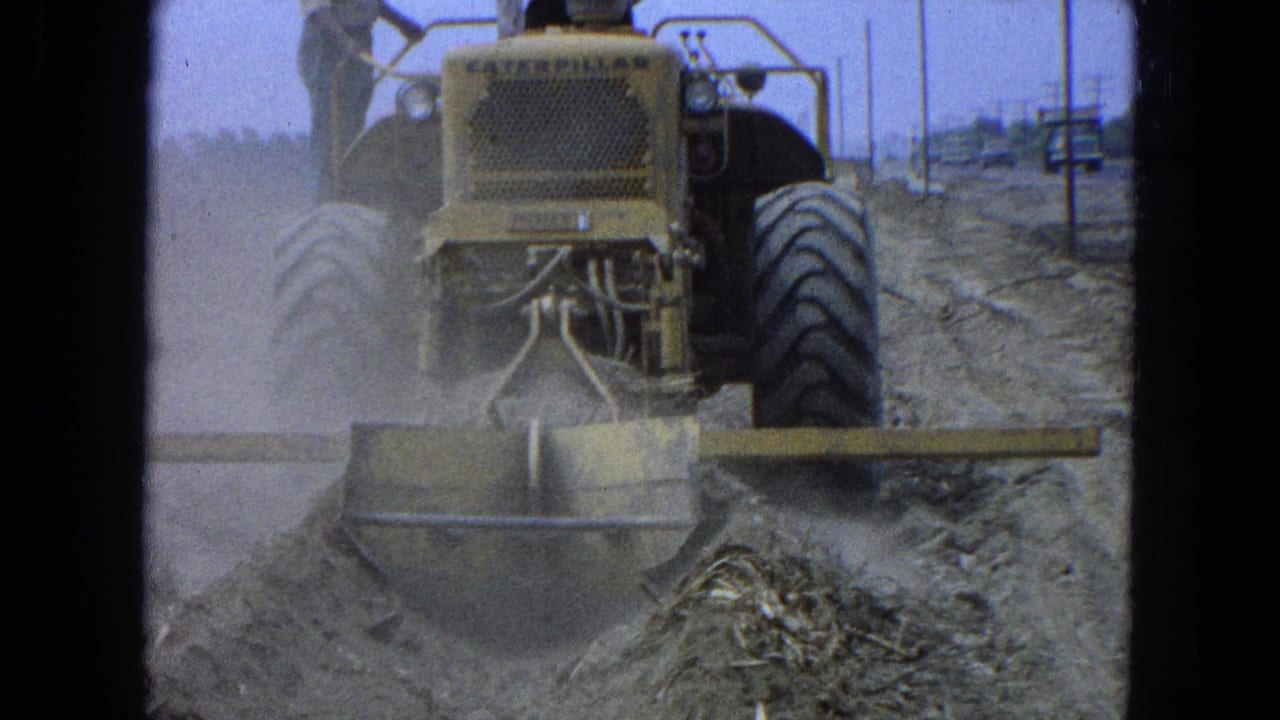Yellow Caterpillar bulldozer grading a dusty dirt worksite with a worker standing on the rear.