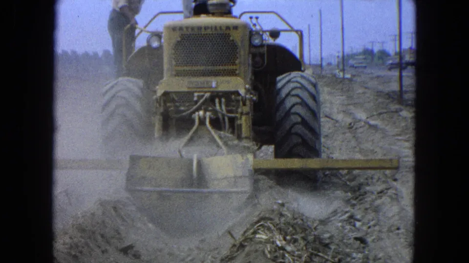 1960s 8mm frame of a Caterpillar bulldozer at a dusty construction site, with a worker standing on the rear platform.