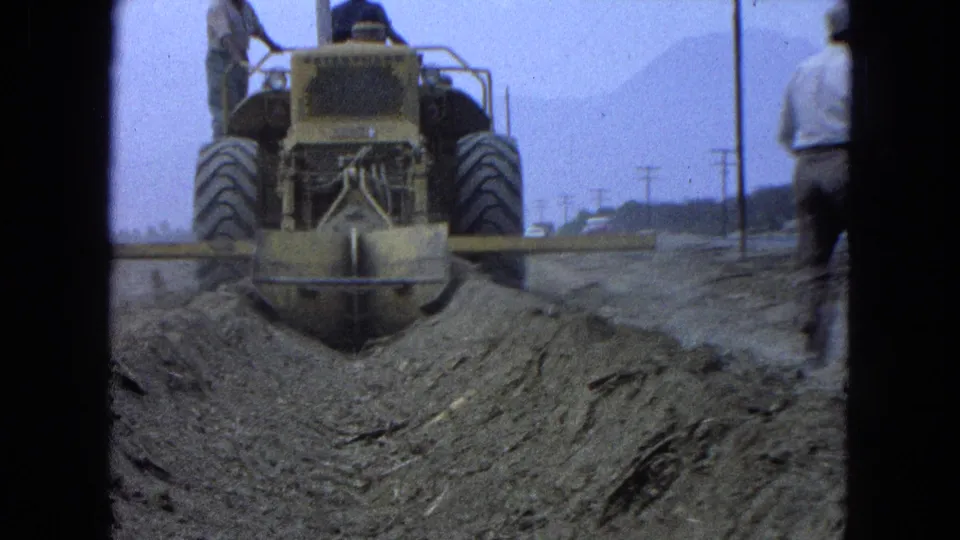 Pueblo, Mexico 1965 home-movie frame showing a bulldozer moving away on a dirt worksite as an adult man walks nearby.