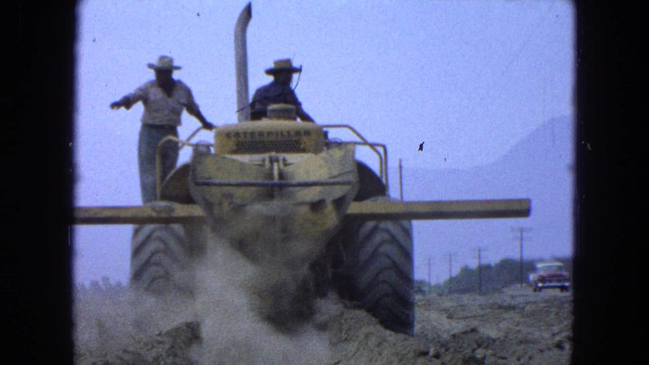 Two men in wide-brimmed hats ride or stand on a yellow earthmover amid dust.