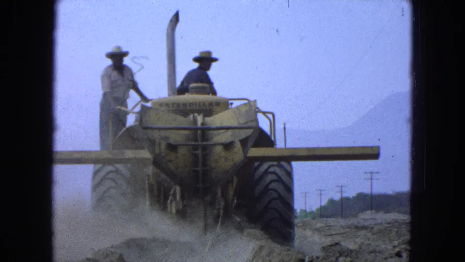 1965 Pueblo, Mexico 8mm frame showing two men on a yellow earthmover with dust and utility poles in the background.