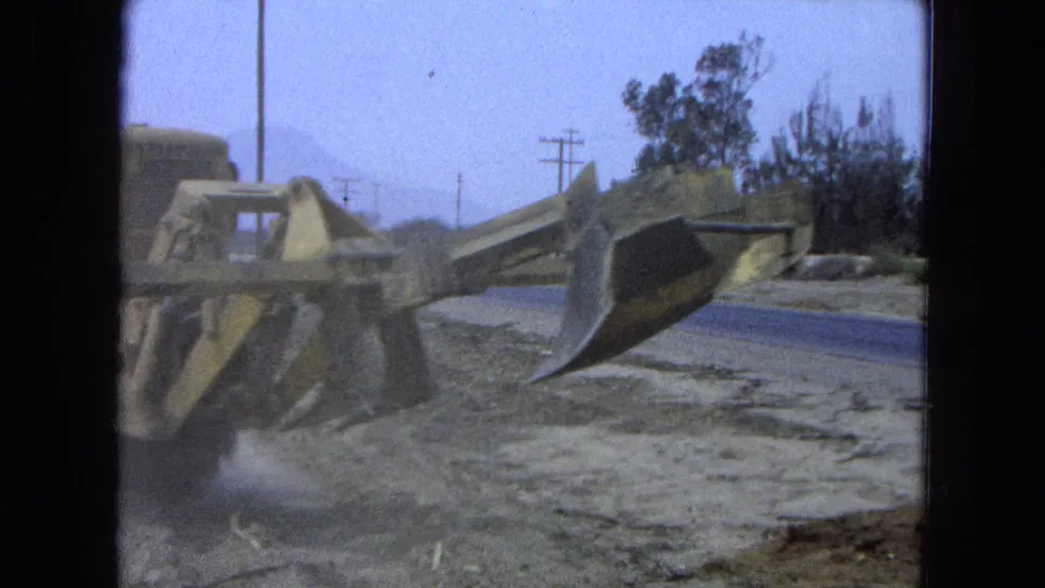 1965 Pueblo, Mexico frame showing the side blade of a yellow earthmover beside a road, with trees and utility poles beyond.